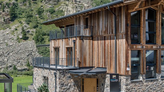 A modern building made of wood and stone in a picturesque landscape. The facade is rustic and blends well into the surroundings. - Gletscherblick - Serviced Apartments - Obergurgl 