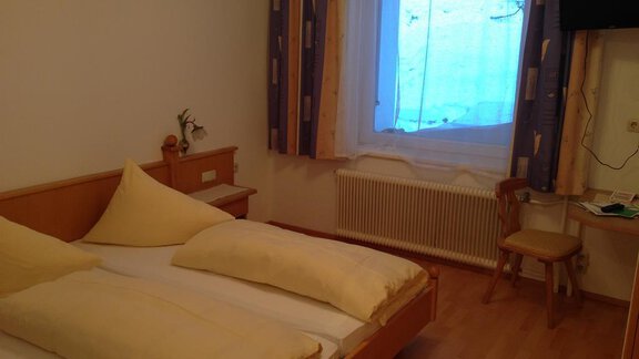 A simple room with two beds, bright bed covers, and a small table. A window with curtains provides daylight. - Haus Timmelseck - Obergurgl 