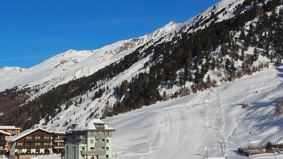 A winter landscape with snow-covered mountains and a clear blue sky. In the foreground, there are some buildings and a ski slope. - Haus Verwall Appartements - Obergurgl 
