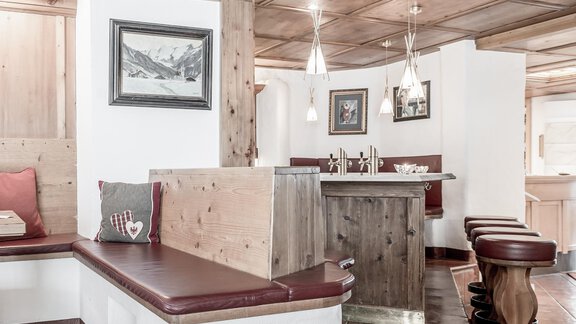 A cozy interior with wooden decorations and comfortable seating. Hanging lamps are at the bar and pictures are on the walls. - Hotel Olympia - Obergurgl 