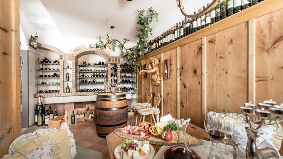 A cozy wine cellar with wooden shelves filled with bottles of wine. On the table, there is a selection of cheese and cold cuts, as well as glasses of red wine. - Hotel Olympia - Obergurgl 