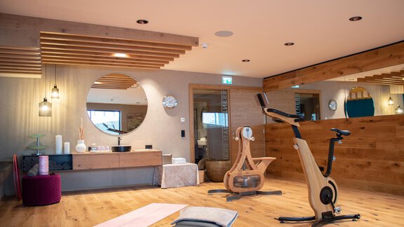 A modern gym with wooden floors and elegant furniture. The room features a fitness machine, a yoga mat, and large mirrors. - Hotel Olympia - Obergurgl 