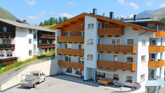 A multi-story building with wooden balconies in a picturesque mountain landscape. In the parking lot, there is a vehicle, and in the background, green hills can be seen. - Haus Martinshof - Obergurgl 
