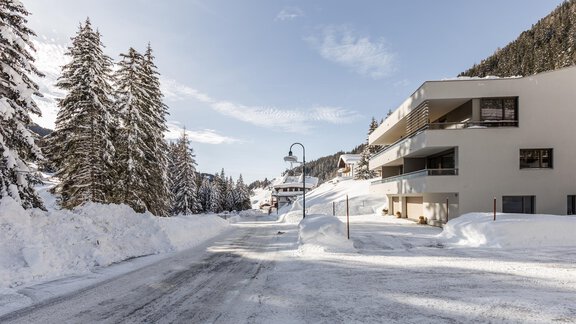 Eine verschneite Straße mit hohen Tannenbäumen und modernen Gebäuden. Der Himmel ist klar und die Landschaft wirkt friedlich. - Apartment Waldesruh - Ochsengarten