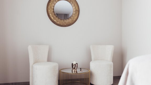Zwei stilvolle, weiße Sessel stehen gegenüber und umgeben einen runden, goldenen Tisch. Ein runder Spiegel hängt an der Wand, was dem Raum eine elegante Note verleiht. - Hotel habicher hof - Oetz