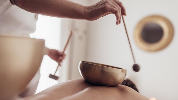 Eine entspannende Wellness-Anwendung mit einer Klangschale auf dem Rücken eines Kunden. Die Therapeutin schlägt sanft auf die Schale, um beruhigende Töne zu erzeugen. - Hotel habicher hof - Oetz