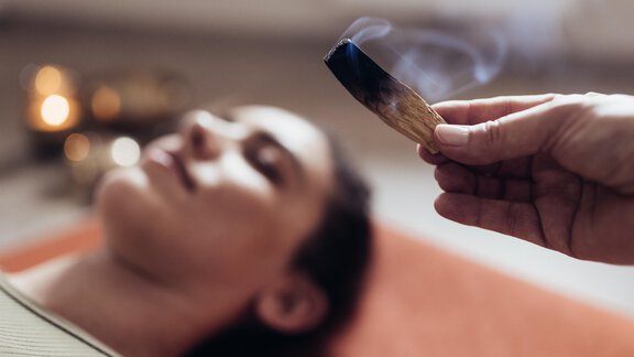 Eine Person liegt entspannt auf einer Matratze, während Räucherwerk über ihr gehalten wird. Der Raum ist gemütlich und sanft beleuchtet. - Hotel habicher hof - Oetz