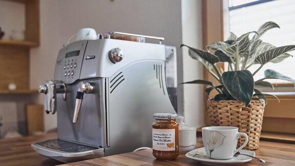 Eine moderne Espressomaschine steht auf einem Holztisch. Neben ihr befindet sich eine Tasse und ein Glas mit Marmelade, umgeben von einer Pflanze. - Adventure Paradise - Sautens