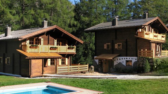 Two cozy wooden block houses in nature, surrounded by trees. A floating pool is visible in the foreground. - Almdorf Waldesruh - Sölden
