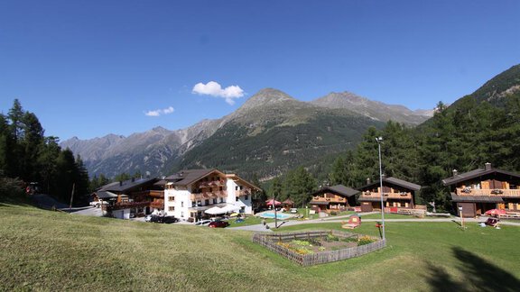 A picturesque alpine landscape with several cozy chalets. Majestic mountains rise in the background under a clear sky. - Almdorf Waldesruh - Sölden