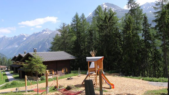 A playground in the mountains with playground equipment and surrounded by trees. Snow-capped mountains and a clear sky are visible in the background. - Almdorf Waldesruh - Sölden