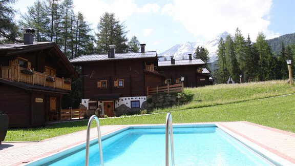 A beautiful outdoor area with a swimming pool and mountain huts in the background. The surroundings are green with trees and a clear blue sky. - Almdorf Waldesruh - Sölden