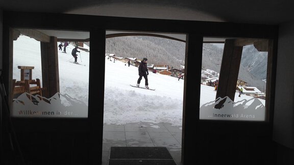 An entrance overlooking a snow-covered slope and skiers. The surroundings showcase typical alpine architecture and snowy mountains. - Am Trogwohl - Sölden