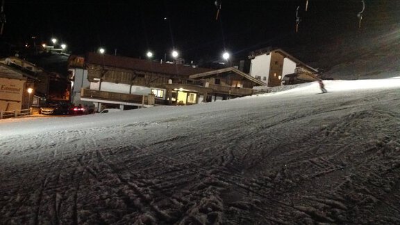 A snowy slope at night with illuminated buildings in the background. A skier is descending the slope. - Am Trogwohl - Sölden