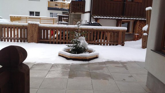 A snow-covered patio with a small fir tree in the center. In the background, wooden fences and buildings can be seen. - Am Trogwohl - Sölden
