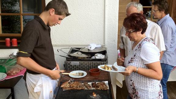 Eine Person grillt Fleisch auf einer Terrasse, während zwei weitere Personen aufmerksam zuschauen. Im Hintergrund sind weitere Menschen und Holzwände sichtbar. - Andis Almdorf - Sölden