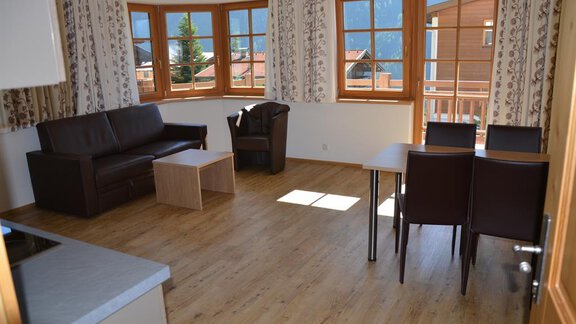 A bright living room with a sofa, a table, and chairs. Large windows provide a view of the surrounding houses and the landscape. - Andis Almdorf - Sölden