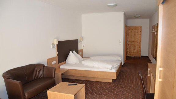 A simple hotel room with two beds, a sofa, and a small table. The room is bright and minimally furnished. - Andis Almdorf - Sölden
