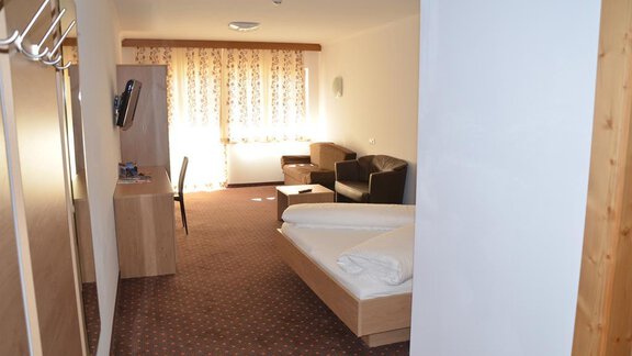A bright hotel room with a double bed, a TV table, and a sofa. Large windows provide plenty of daylight. - Andis Almdorf - Sölden