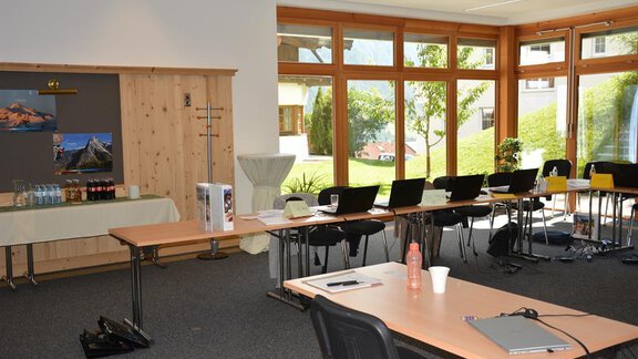 A bright seminar room with large windows and a view of greenery. Tables and chairs are prepared for a meeting. - Andis Almdorf - Sölden