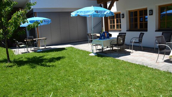 A well-maintained garden with a lawn area and a table with a sun umbrella. Several chairs are arranged around the table, next to a light-colored building. - Andis Almdorf - Sölden