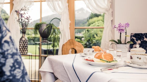 A beautifully set table with rolls and fruit. In the background are large windows overlooking green meadows and mountains. - Andre Arnold - Boutique Hotel - Sölden