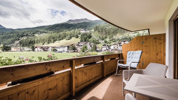 A balcony with a wooden railing and chairs, from which one has a view of green meadows and mountains. The sky is partly cloudy. - Andre Arnold - Boutique Hotel - Sölden