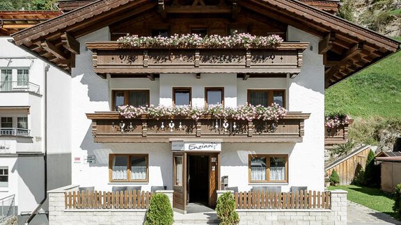 A traditional house in the mountains with blooming balcony boxes. The surroundings feature green grass and trees in the background. - Apart Garni Enzian - Sölden | © RudiWyhlidal