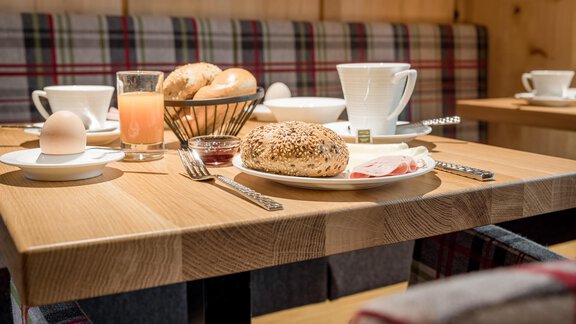 A cozy breakfast table with rolls, an egg, a glass of juice, and cups. In the background, there are benches with a checkered design. - Apart Garni Enzian - Sölden