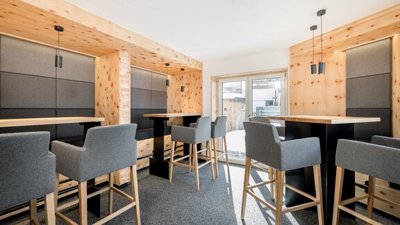 A modern café with tall tables and chairs. The walls are made of light wood, and plenty of daylight streams through the large windows. - Apart Garni Enzian - Sölden