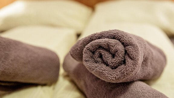 A rolled-up towel in brown is lying on a bed with light pillows. The scene conveys a sense of relaxation and coziness. - Apart Garni Enzian - Sölden