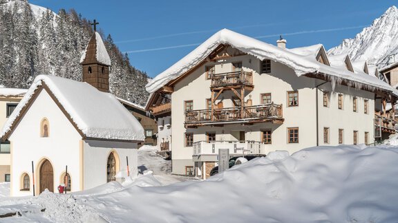 A picturesque winter landscape with snow-covered cottages and a small church. In the background, majestic mountains rise under a clear blue sky. - Apart Hotel Garni Alt Kaisers - Sölden
