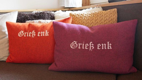 Zwei dekorative Kissen in Rot und Lila mit dem Schriftzug "Grieß enk". Sie sind auf einem Sofa platziert und verleihen dem Raum eine gemütliche Atmosphäre. - Alpen Diamond Top 100 | 300 | 700 - Sölden