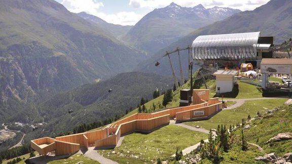 An impressive mountain landscape with green meadows and majestic peaks. In the foreground, there is a modern cable car and a wooden walkway. - Appartement  Fabio - Sölden
