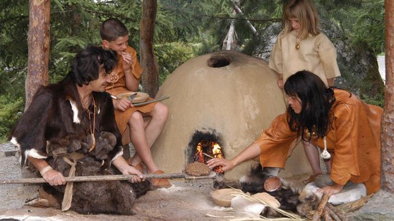 A group of prehistoric people sits around a small clay oven. They are dressed in simple garments and are engaged in preparing food. - Appartement  Fabio - Sölden