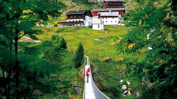 A suspension bridge leads through a green landscape to a picturesque farmhouse. The surroundings are surrounded by trees and mountains. - Appartement  Fabio - Sölden