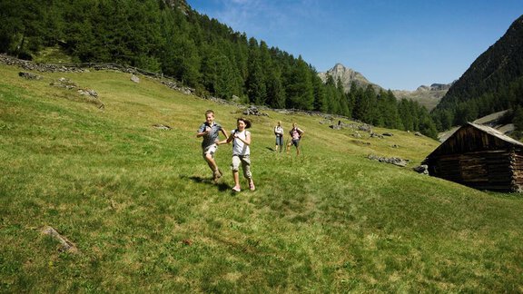 Children are happily running across a green meadow in the mountains. In the background, there are trees and a small wooden house. - Appartement  Fabio - Sölden