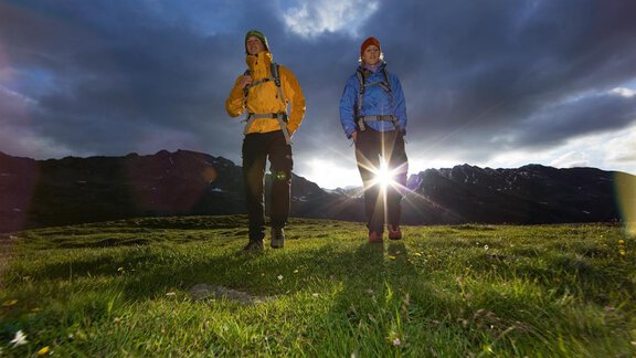 Two hikers stand on a green meadow in front of a dramatic cloud backdrop. The sun shines behind them, illuminating the scene. - Appartement  Fabio - Sölden