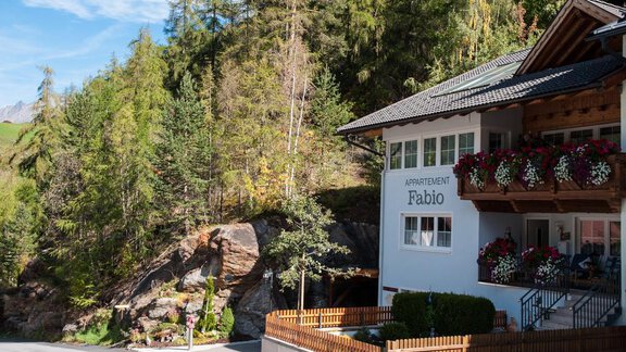 A cozy apartment building named "Fabio" is situated in a natural setting. The facade is adorned with flowers, and in the background, green trees and a blue sky can be seen. - Appartement  Fabio - Sölden
