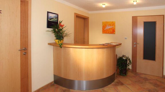 A bright reception room with a round wooden reception desk. Pictures hang on the wall, and a plant adds color to the room. - Appartement Rangger - Sölden