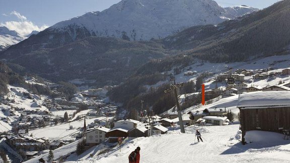 A snowy mountain landscape with skiers on a slope. In the background, high mountains and a picturesque village can be seen. - Appartement Rangger - Sölden