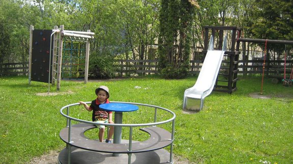 A child is playing on a carousel at a playground. In the background, a slide and a climbing frame can be seen. - Appartement Rangger - Sölden