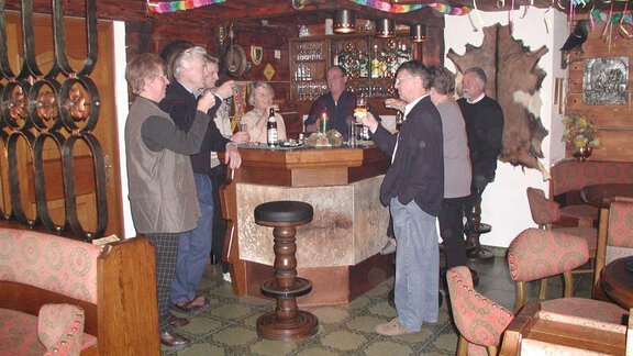 A group of people stands at a bar and toasts each other. The room has a cozy atmosphere with wooden paneling and comfortable seating. - Appartementhaus Karlinger - Sölden