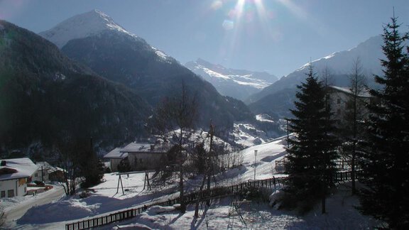 A snowy mountain landscape with bright sunshine. In the foreground, some houses and trees are visible. - Appartementhaus Karlinger - Sölden