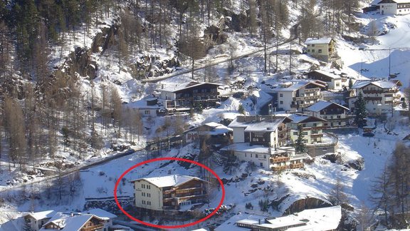 A snowy landscape with several chalets and a highlighted building in the center. The surroundings are surrounded by fir trees and mountains. - Appartements Waldesrand - Sölden