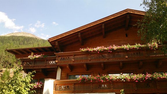 A traditional wooden chalet with a balcony and blooming plants. In the background, green mountains are visible under a blue sky. - Appartements Waldesrand - Sölden