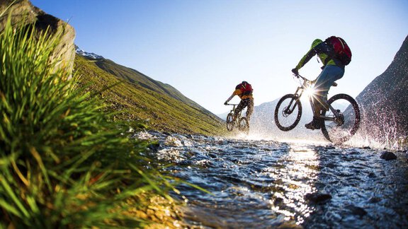 Two cyclists ride over a splashing brook in a sunny landscape. In the background, green hills and a clear blue sky can be seen. - Appt. Gisela - Sölden