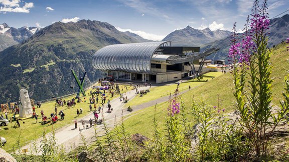 A modern mountain station with a glass facade amidst a picturesque mountain landscape. In the foreground, colorful flowers are in bloom and people are enjoying the view. - Appt. Gisela - Sölden