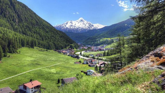 A picturesque mountain landscape with snow-capped peaks and a green valley. In the foreground, there are small houses and a peaceful town. - Appt. Gisela - Sölden