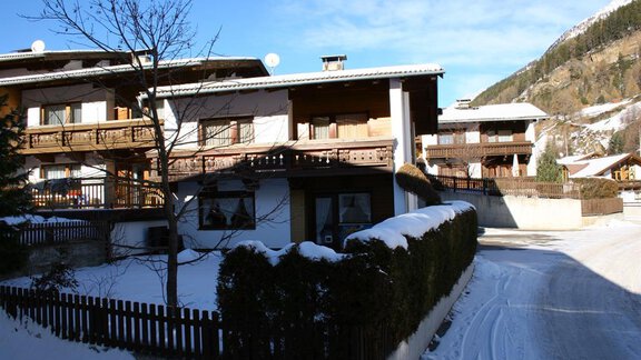 A picturesque mountain village with snow-covered houses and a quiet street. The surroundings are filled with gentle hills and trees. - Appt. Schnöller - Sölden